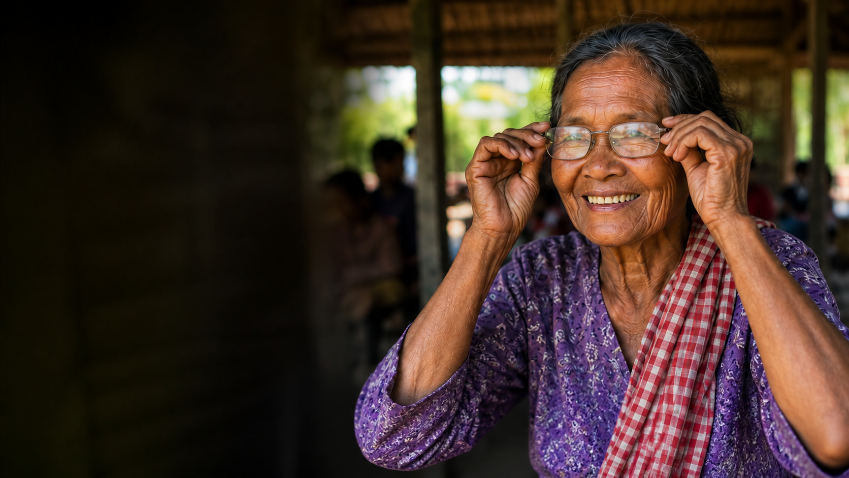 An older Cambodian woman smiling as she puts on reading glasses